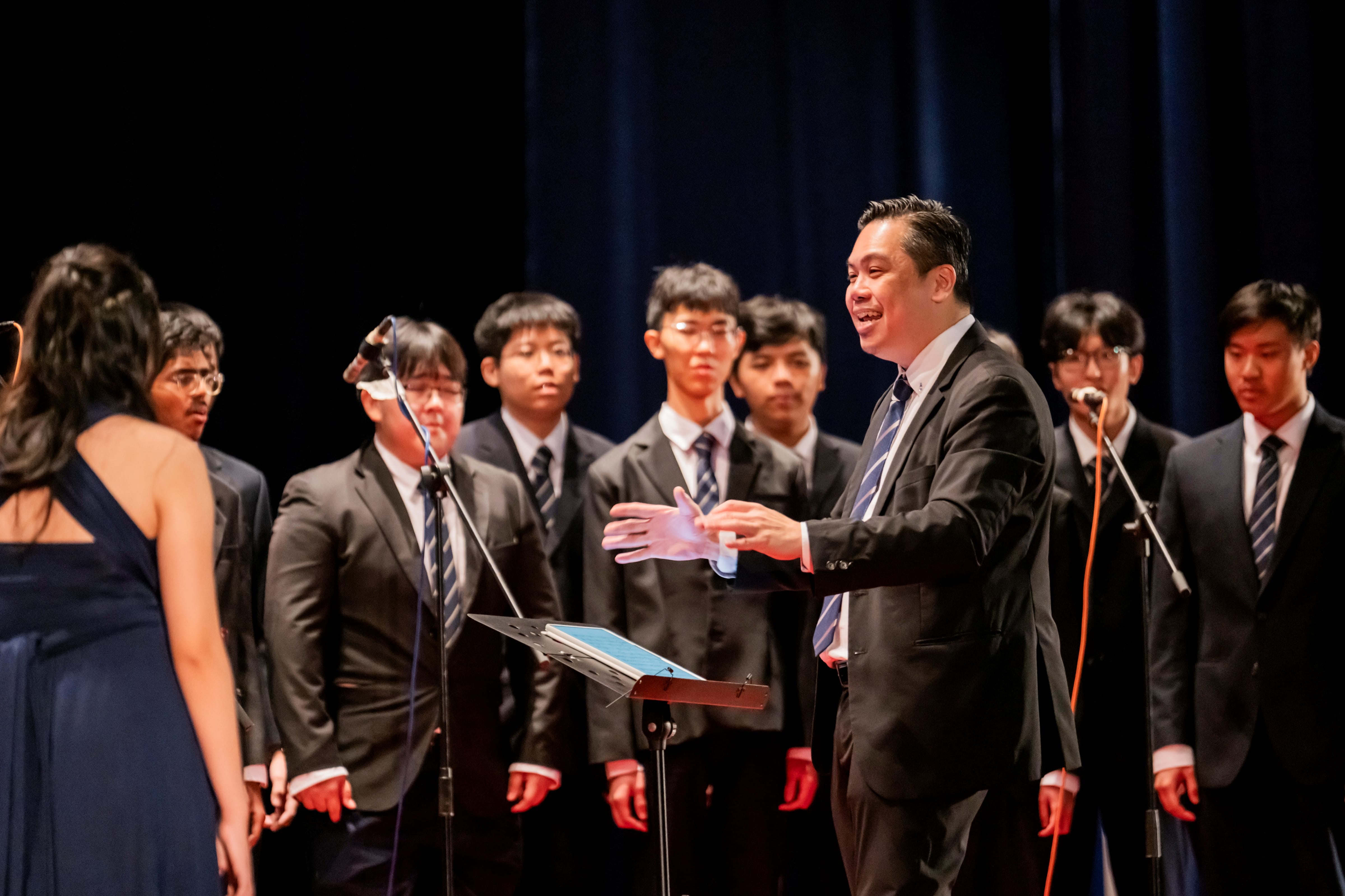 A choir conductor gesturing with open hands as he directs student singers on stage during a musical performance.